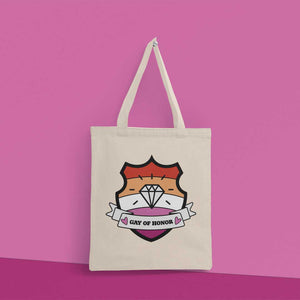 Natural coloured tote bag hanging from a peg on a pink wall. The bag features a shield with a banner across it's front. Banner reads Gay of Honor. The background of the shield is the lesbian pride flag and there is a jewel icon in the centre.