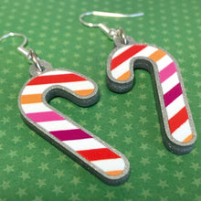 Load image into Gallery viewer, Silver glitter candy cane dangle earrings lay on a green star paper background. The stripes on the candy cane are those of the Lesbian flag; orange, pink, and white. One of the earrings is turned over to show the reverse, which is plain silver glitter.