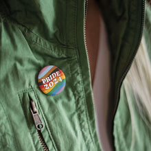 Load image into Gallery viewer, Close up of a person wearing a green zip up jacket with a 38mm badge pinned to it. The badge features the colours of the progress pride flag in waving lines across the background with retro style white typography in the foreground reading Pride 2024