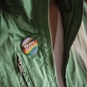 Close up of a person wearing a green zip up jacket with a 38mm badge pinned to it. The badge features the colours of the progress pride flag in waving lines across the background with retro style white typography in the foreground reading Pride 2024
