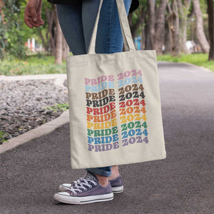 Close up of a person holding a natural coloured tote bag in a park setting. Print on the bag is retro style typography reading Pride 2024 repeated 10 times, each line is a different colour of the Progress Pride Flag.