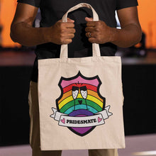 Load image into Gallery viewer, Close up of a black person holding a natural coloured tote bag featuring a shield with a banner across it&#39;s front. Banner reads Pridesmate. The background of the shield is the Gilbert Baker pride flag and there is a pair of clinking champagne flutes in the centre.