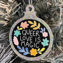 Load image into Gallery viewer, Close up of a white Christmas tree displaying a silver glitter bauble featuring the text  Queer Love is Magic and hand drawn illustrations of flowers in rainbow colours.