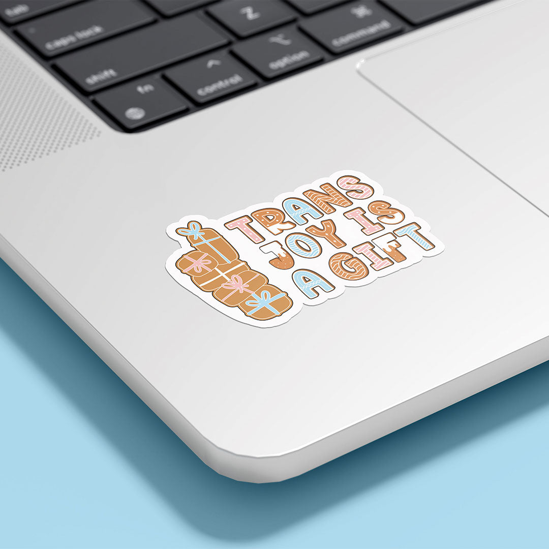 A "Trans Joy Is A Gift" sticker featuring cute gingerbread presents, attached to the corner of a silver laptop on a light blue background.
