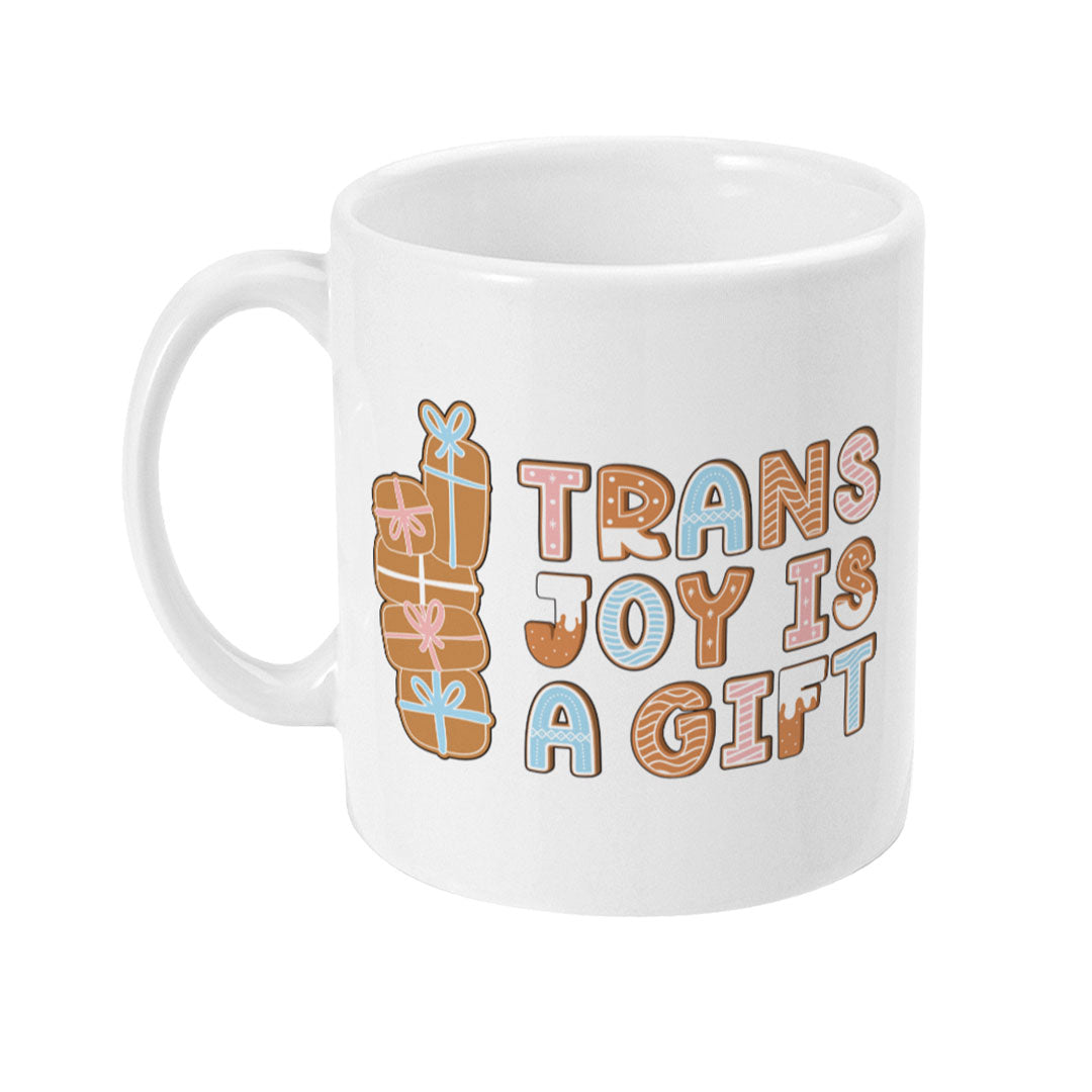 Left side view of a white mug featuring the text "Trans Joy is a Gift" in gingerbread-style lettering, with stacked presents in pink, blue, and gingerbread brown on the side.