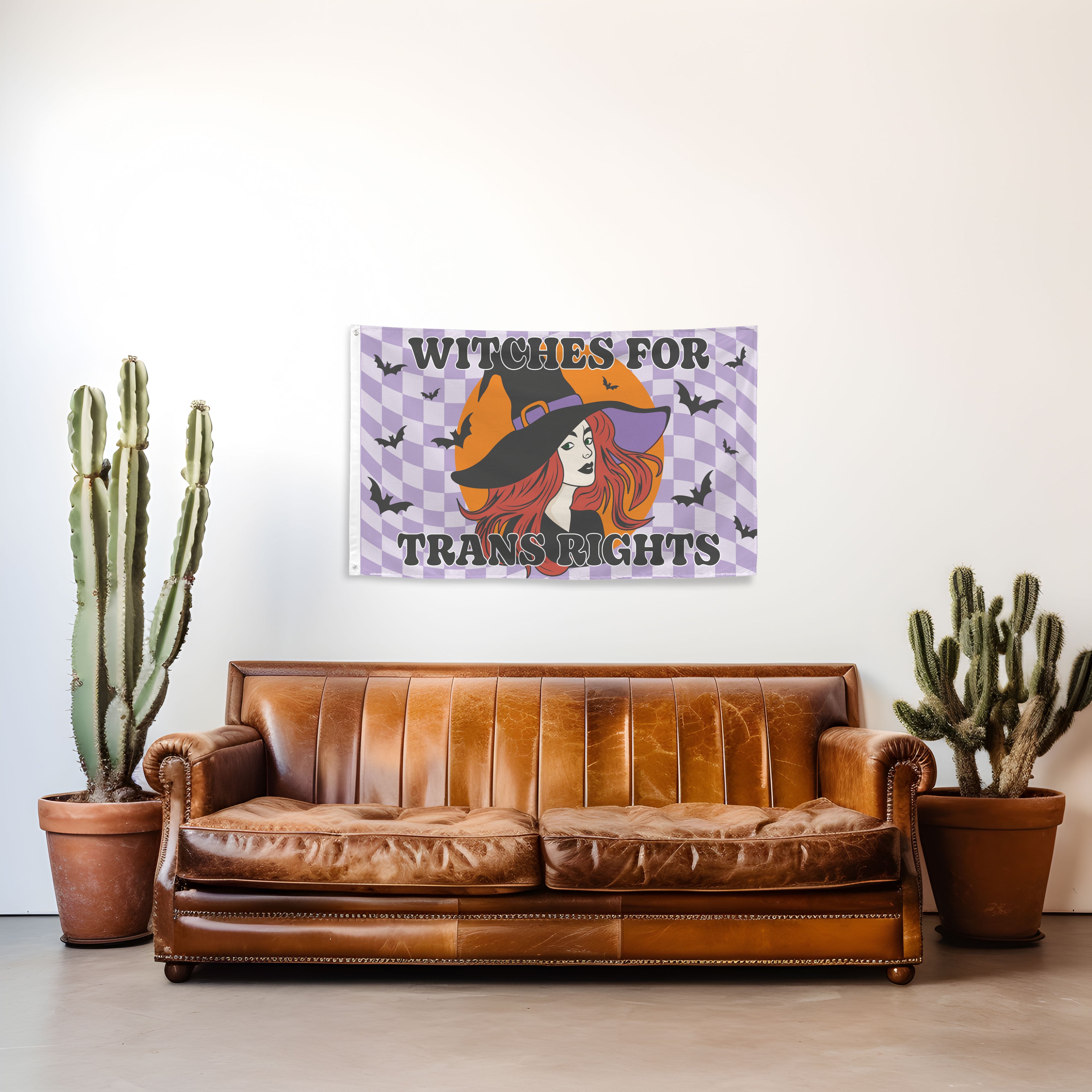 Brown leather sofa with a 'Witches for Trans Rights' poster on a white wall, flanked by two potted cacti.