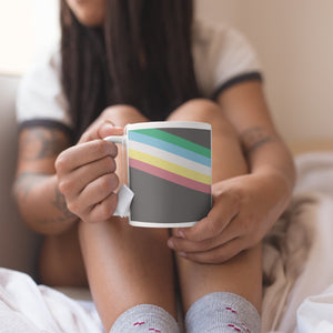 Person sat on their bed holding a ceramic coffee mug featuring the disabled pride flag.
