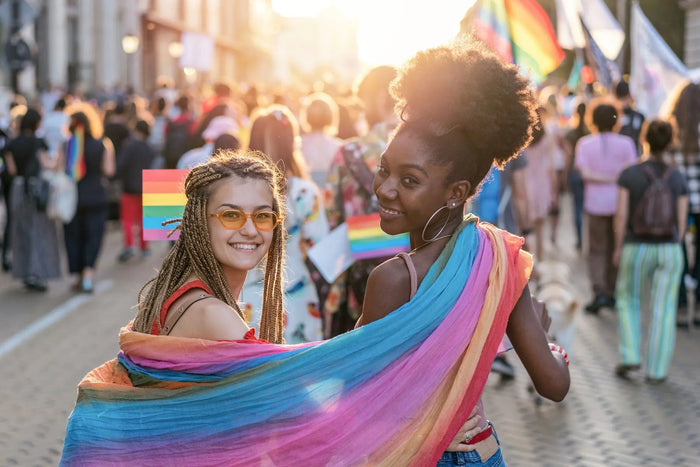 Two young women with a rainbow scarf draped around them ready to march in a pride parade.