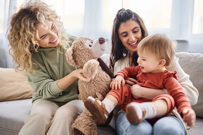 A loving same-sex couple sits on a cosy sofa with their young child, sharing a joyful moment. One parent, wearing a cream jumper, holds the child securely, while the other, dressed in a green hoodie, plays with a large teddy bear. The child, dressed in a 