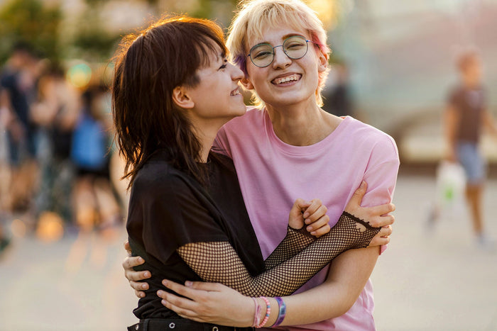 A joyful moment between two people embracing each other outdoors. One person, wearing a black mesh-sleeved top, smiles warmly while leaning into the other, who is dressed in a light pink t-shirt and round glasses. They share a tender connection, with both