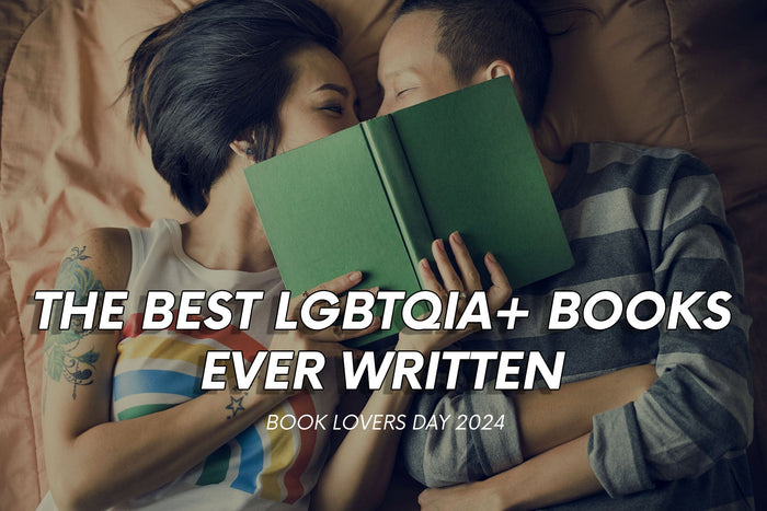 Text reads 'The Best LGBTQIA+ Books Ever Written - Book Lovers Day 2024'. In the background is an image of a lesbian couple lying on a bed holding a book  slightly covering their faces.