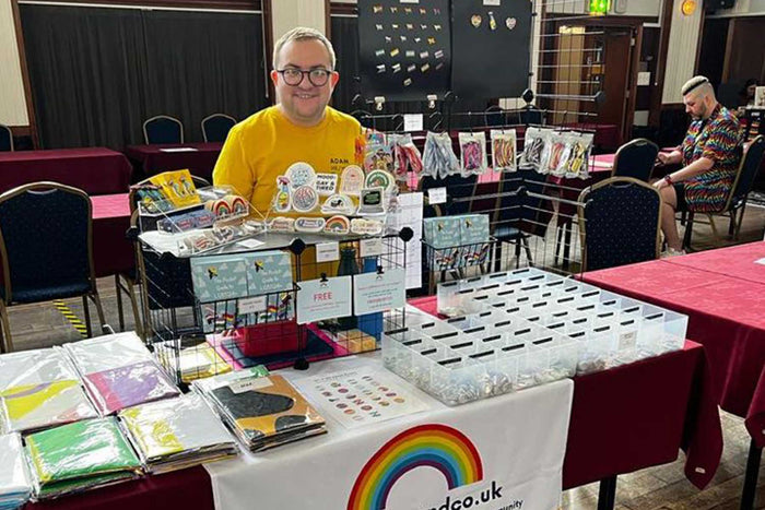 Adam stands behind the Rainbow & Co stall at Bury Pride 2023 which is full of LGBTQIA+ goodies including badges, stickers, flags, enamel pins, and shoelaces.