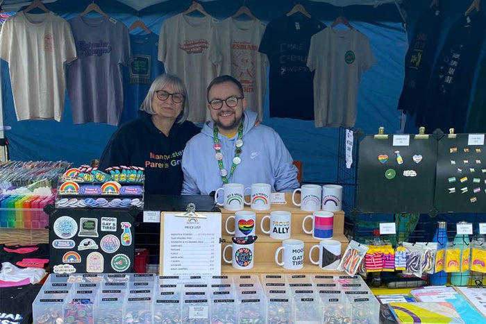 Adam and his Mum stand behind the Rainbow & Co stall at Durham Pride 2023 which is full of LGBTQIA+ goodies including badges, stickers, flags, enamel pins, and shoelaces.