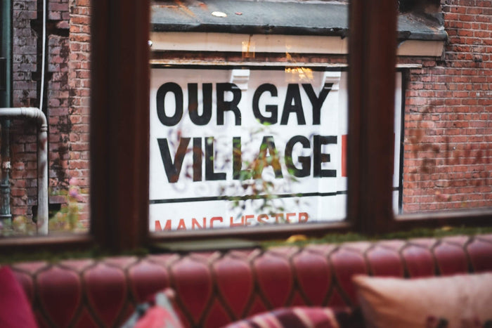 View out of the window of a bar on Canal St, Manchester, showing a piece of wall art designed to look like a street sign with the text OUR GAY VILLAGE