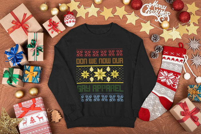  A black festive sweatshirt with the text “Don We Now Our Gay Apparel” in colourful lettering, surrounded by a holiday-themed knit-style pattern of snowflakes and stars, displayed on a wooden background with Christmas decorations.