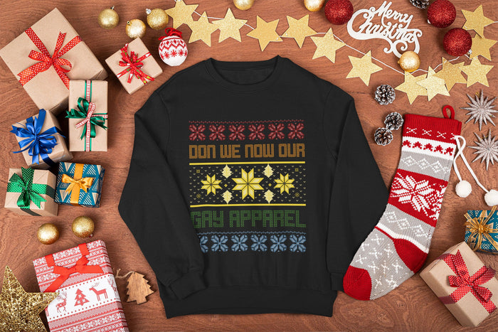  A black festive sweatshirt with the text “Don We Now Our Gay Apparel” in colourful lettering, surrounded by a holiday-themed knit-style pattern of snowflakes and stars, displayed on a wooden background with Christmas decorations.