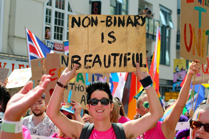 A non binary person marching in a pride parade holding a placard reading Non Binary is Beautiful