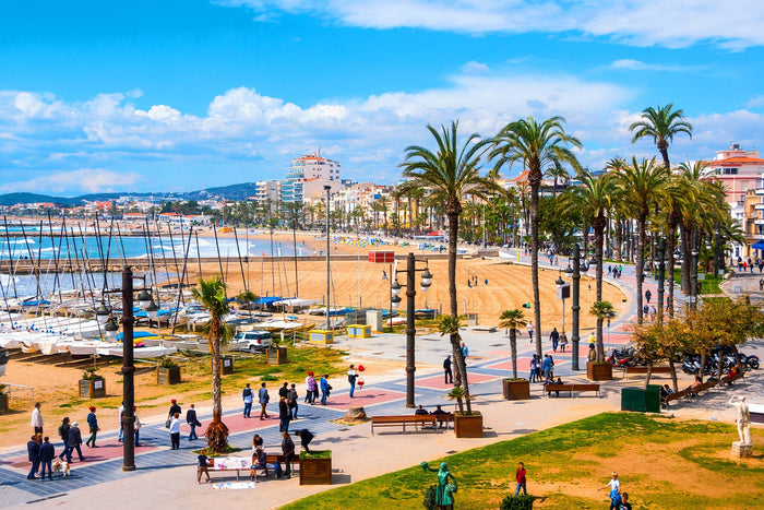 A sunny day in Sitges, Spain, showing the palm-lined promenade, golden sandy beach, and people walking along the seafront. Sailboats are docked in the marina, and colourful buildings line the background under a blue sky with scattered clouds.