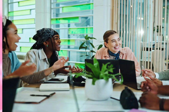 A diverse group of young professionals is engaged in a lively discussion at a modern office table. Two women at the forefront are smiling and laughing, one wearing a bandana and a denim jacket, and the other in a peach blouse with a scarf. The setting is 