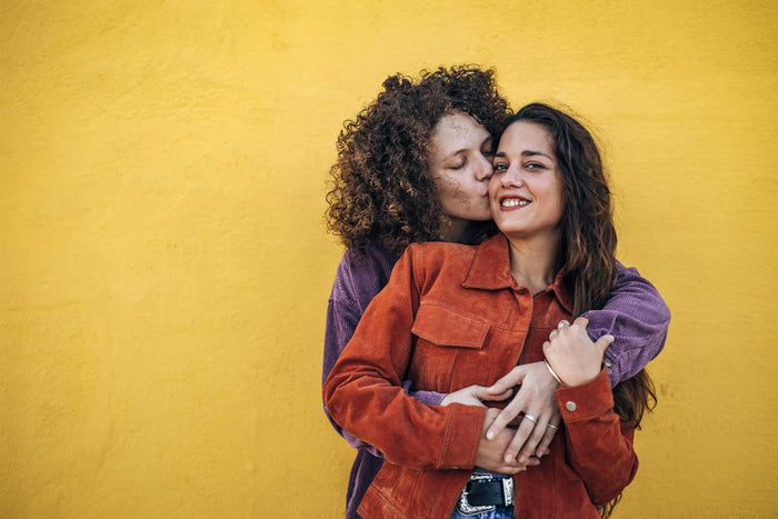 Lesbian couple stood against a yellow wall. One woman is kissing her girlfriend on the cheek.