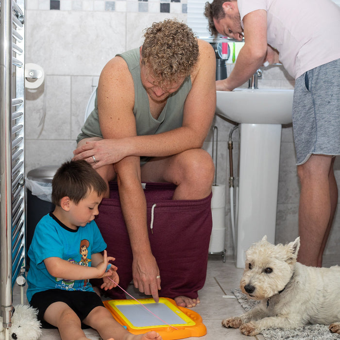 Image shows busy family life in a small bathroom. One dad stands and brushes his teeth whilst the other is sat on the toilet leaning down to entertain their toddler who is sat between his legs on the floor. Joining them is their Westie dog who is also lay