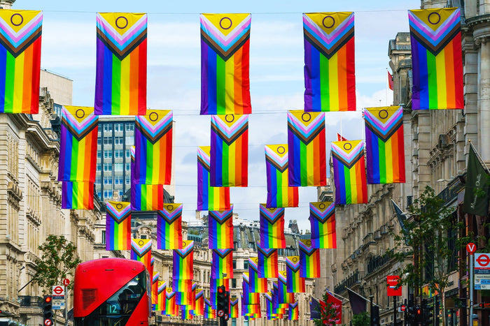 Dozens of Progress Pride flags, including the intersex-inclusive version, hang across a busy London street lined with historic buildings. A red double-decker bus and classic Underground signs are visible, celebrating LGBTQ+ Pride in the heart of the UK ca