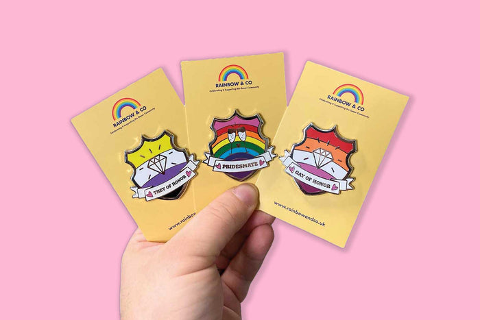 Person holding three enamel pins on yellow backing cards. The pins are each shaped like a shield and carry text reading; They of Honor, Pridesmate, and Gay of Honor.