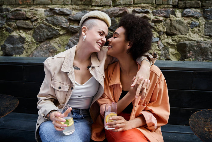 Two sapphic individuals share a joyful and intimate moment, sitting close together outdoors. They are holding drinks with slices of lemon and lime, smiling affectionately at one another, showcasing the beauty and warmth of sapphic love.