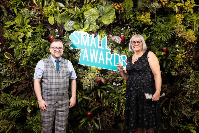 On the left of the image is Adam, wearing a check grey waistcoat and trousers with a pale blue shirt and teal tie. On the right is Adam's Mum Louise, she is wearing a black dress with flowers embroidered on it. They are stood in front of a living wall whi