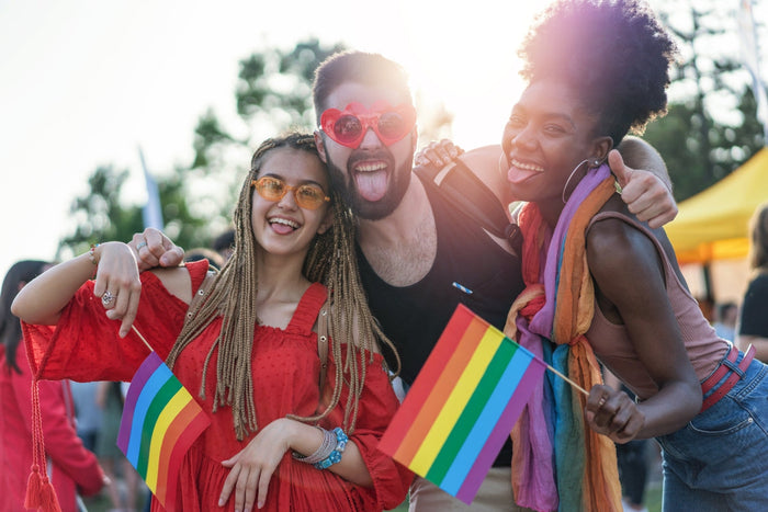 Three people celebrating at a pride event. They are each holding rainbow flags and waving towards the camera pulling silly faces with their tongues out.