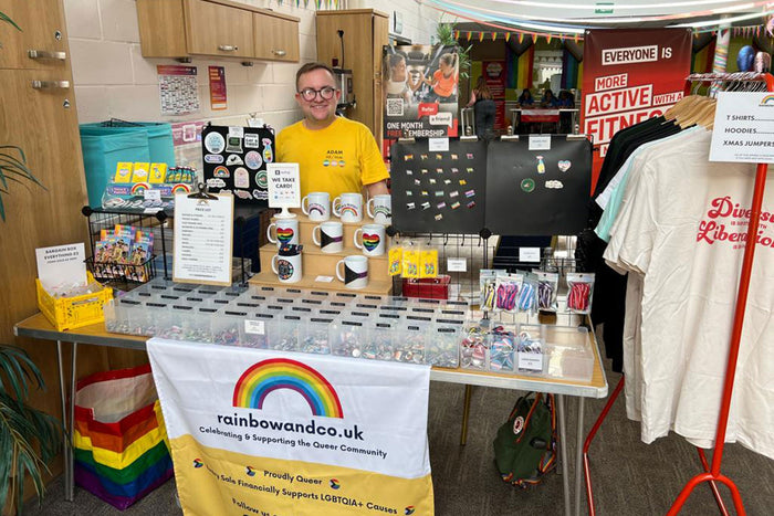 Adam stood behind the Rainbow & Co stall at Wythenshawe Pride 2023, on the stall you can see pride mugs, badges, stickers and pins.