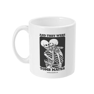 A white ceramic coffee mug with the handle on the left. The mug shows two skeletons embracing and the text 'And They Were Tomb Mates'