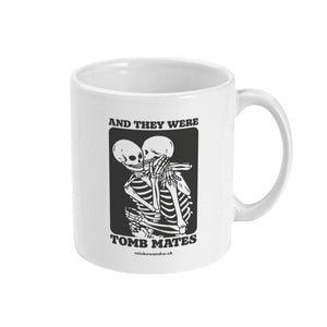 A white ceramic coffee mug with the handle on the right. The mug shows two skeletons embracing and the text 'And They Were Tomb Mates'