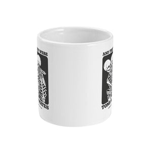 A white ceramic coffee mug with handle facing away from view. The mug shows two skeletons embracing and the text 'And They Were Tomb Mates'