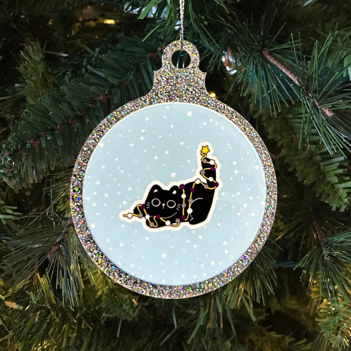 A glittery Christmas bauble showcasing a black cat wrapped in fairy lights in the colours of the asexual pride flag. The cat, adorned with a star on its tail, sits against a snowy background on a festive tree.