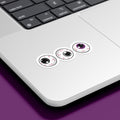 Three circular stickers with eyeball designs are placed on the bottom corner of a silver laptop. The irises are black, grey, and purple, representing the asexual pride flag.
