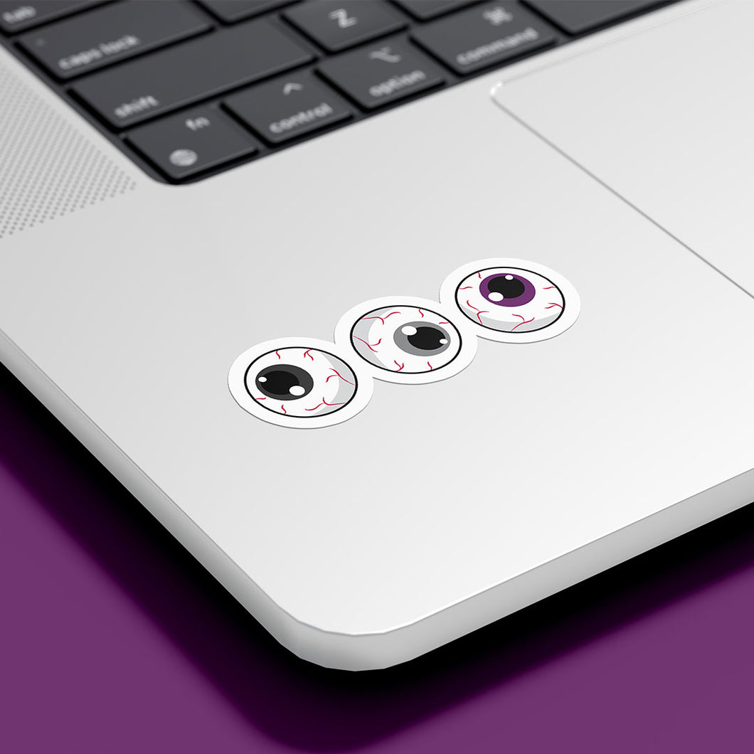 Three circular stickers with eyeball designs are placed on the bottom corner of a silver laptop. The irises are black, grey, and purple, representing the asexual pride flag.