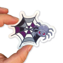 Load image into Gallery viewer, A hand holding a cute spider sticker with an asexual pride web design. The web is coloured in shades of grey, white, black, and purple, representing the asexual flag.