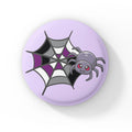 A circular badge featuring a cute grey spider with pink eyes on a web coloured in black, grey, white, and purple, representing the asexual pride flag. The badge has a pastel purple background.