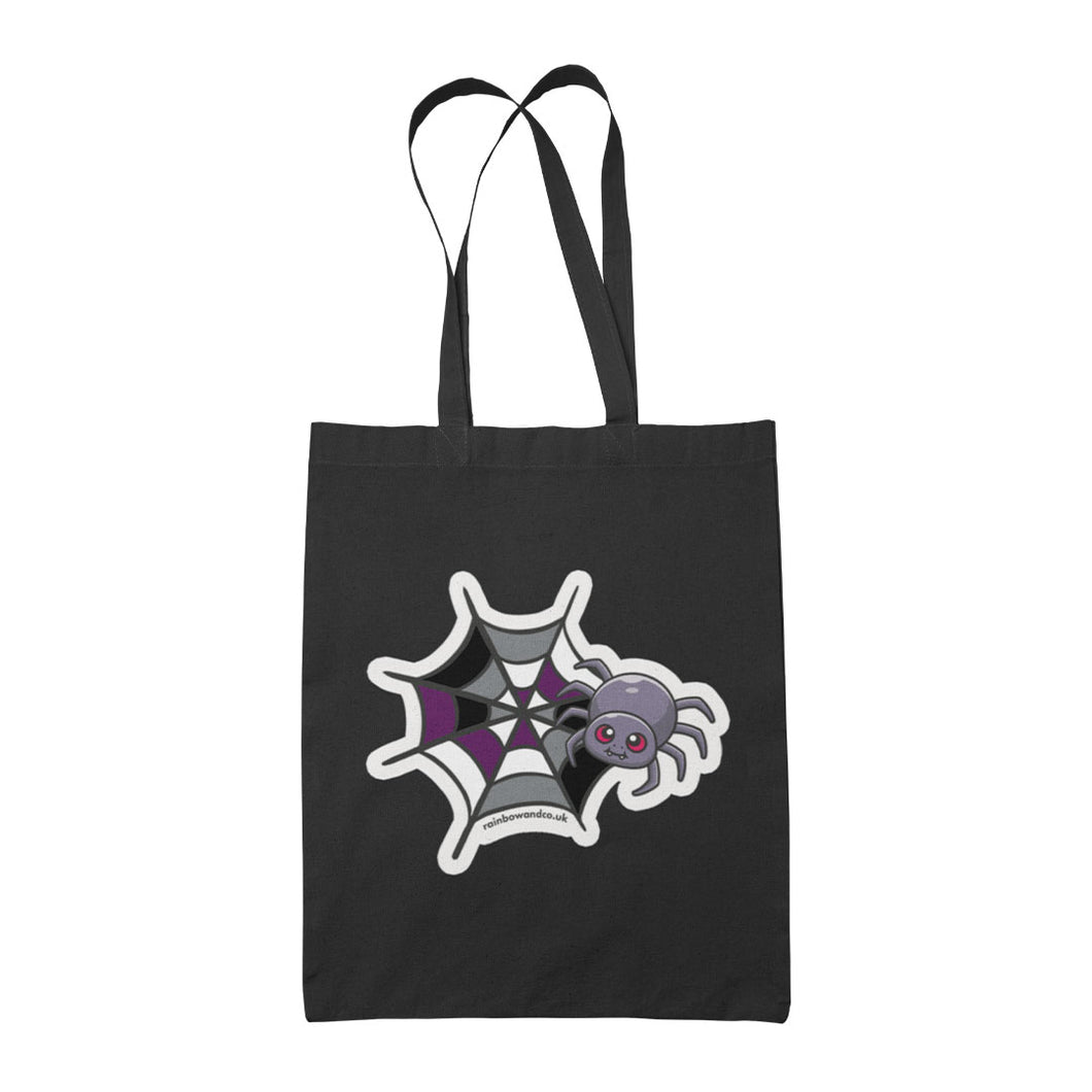 A black tote bag featuring a cute cartoon spider in front of a spider web coloured in the asexual pride flag colours: black, grey, white, and purple.