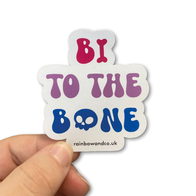 A hand holding a gloss sticker featuring the slogan 'Bi To The Bone' in the colours of the bisexual flag with a bone and a skull icon.