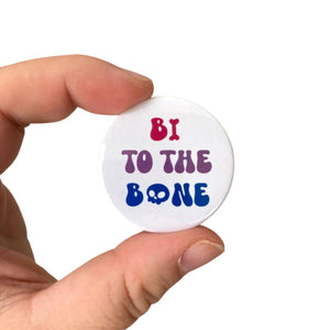 A white 38mm Badge with text reading 'Bi To The Bone' on the colours of the bisexual flag