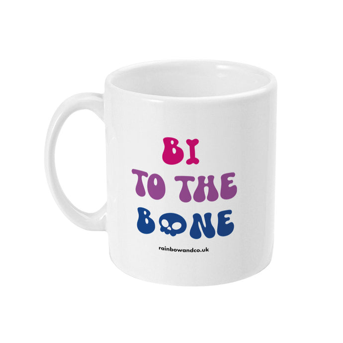 A white ceramic coffee mug with the handle on the left. The mug shows the slogan 'Bi To The Bone' in the colours of the bisexual flag with a bone and a skull icon.