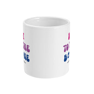 A white ceramic coffee mug with handle facing away from view. The mug shows the slogan 'Bi To The Bone' in the colours of the bisexual flag with a bone and a skull icon.