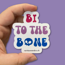 Load image into Gallery viewer, A hand holding a gloss sticker between the thumb and forefinger. The sticker features the slogan &#39;Bi To The Bone&#39; in the colours of the bisexual flag with a bone and a skull icon.