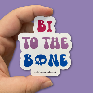A hand holding a gloss sticker between the thumb and forefinger. The sticker features the slogan 'Bi To The Bone' in the colours of the bisexual flag with a bone and a skull icon.