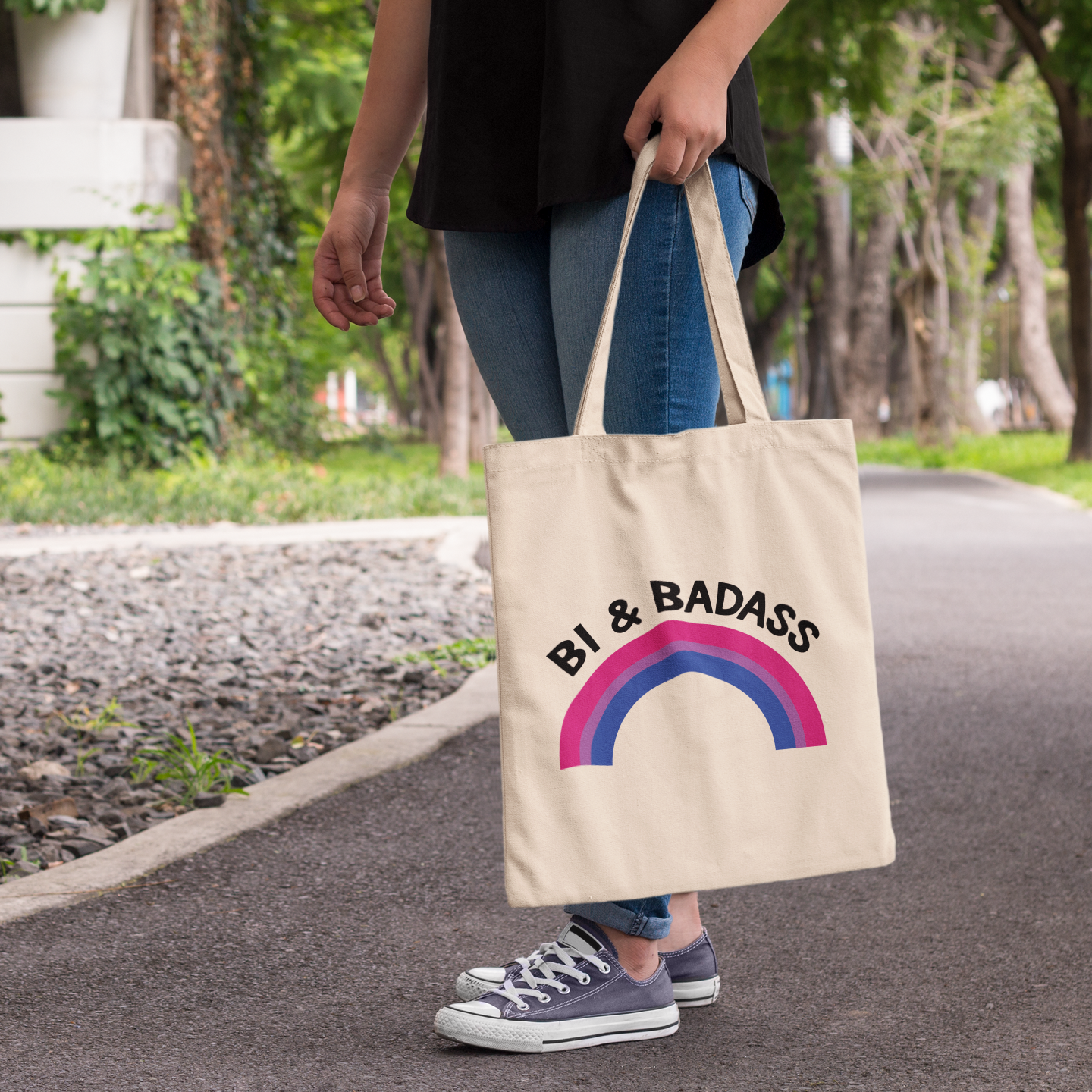Natural cotton tote bag featuring the text “Bi & Badass” with a rainbow arc in bisexual pride colours, worn over the shoulder outdoors