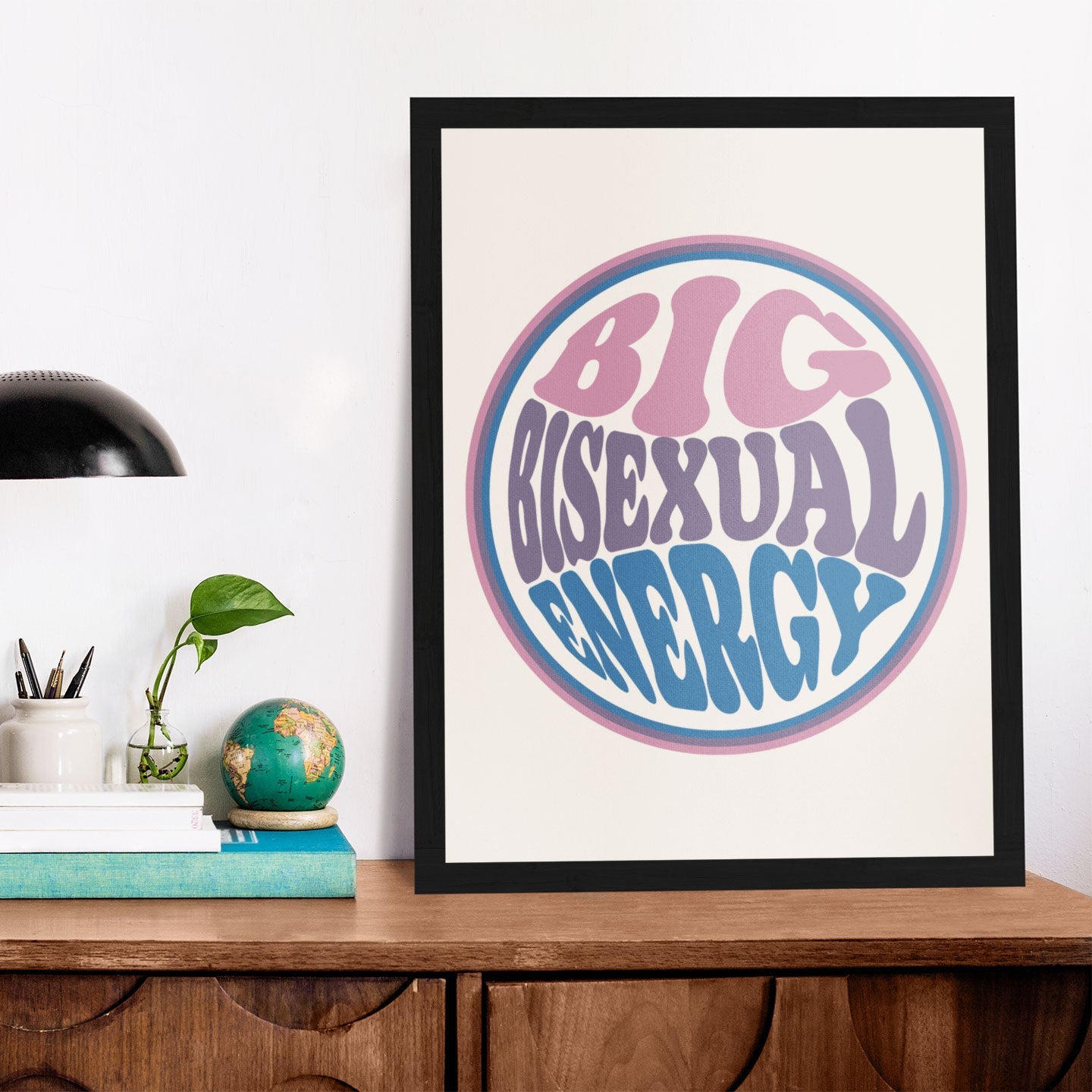 A framed Big Bisexual Energy print in pastel pink, purple, and blue, placed on a wooden desk beside a lamp and books.