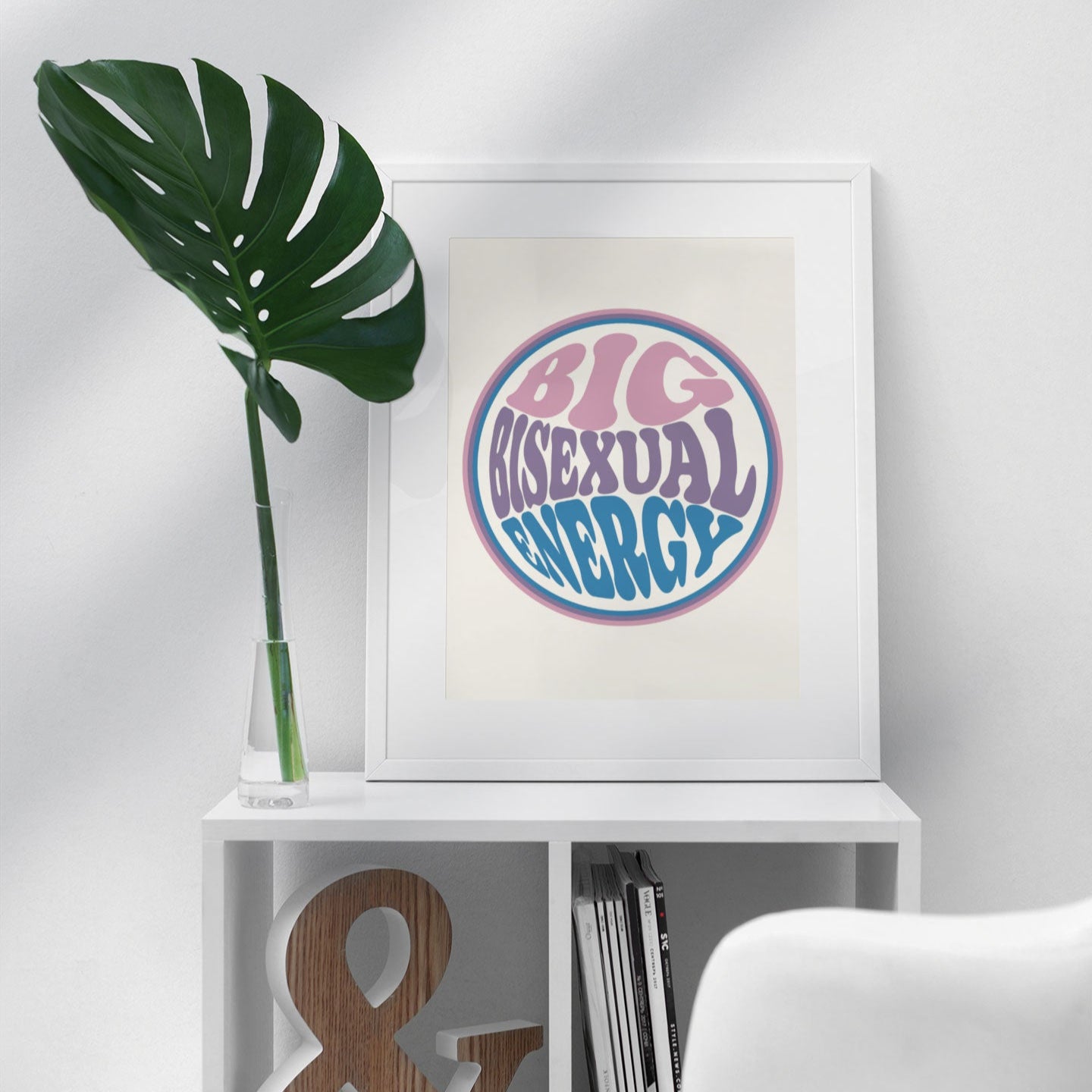 A Big Bisexual Energy print in a modern, minimalist setting, framed in white and styled with a large green monstera plant.