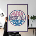A person in black clothing sits on a modern chair, holding a large framed Big Bisexual Energy print in front of their face. The background includes a minimalist desk with a lamp and a plant.